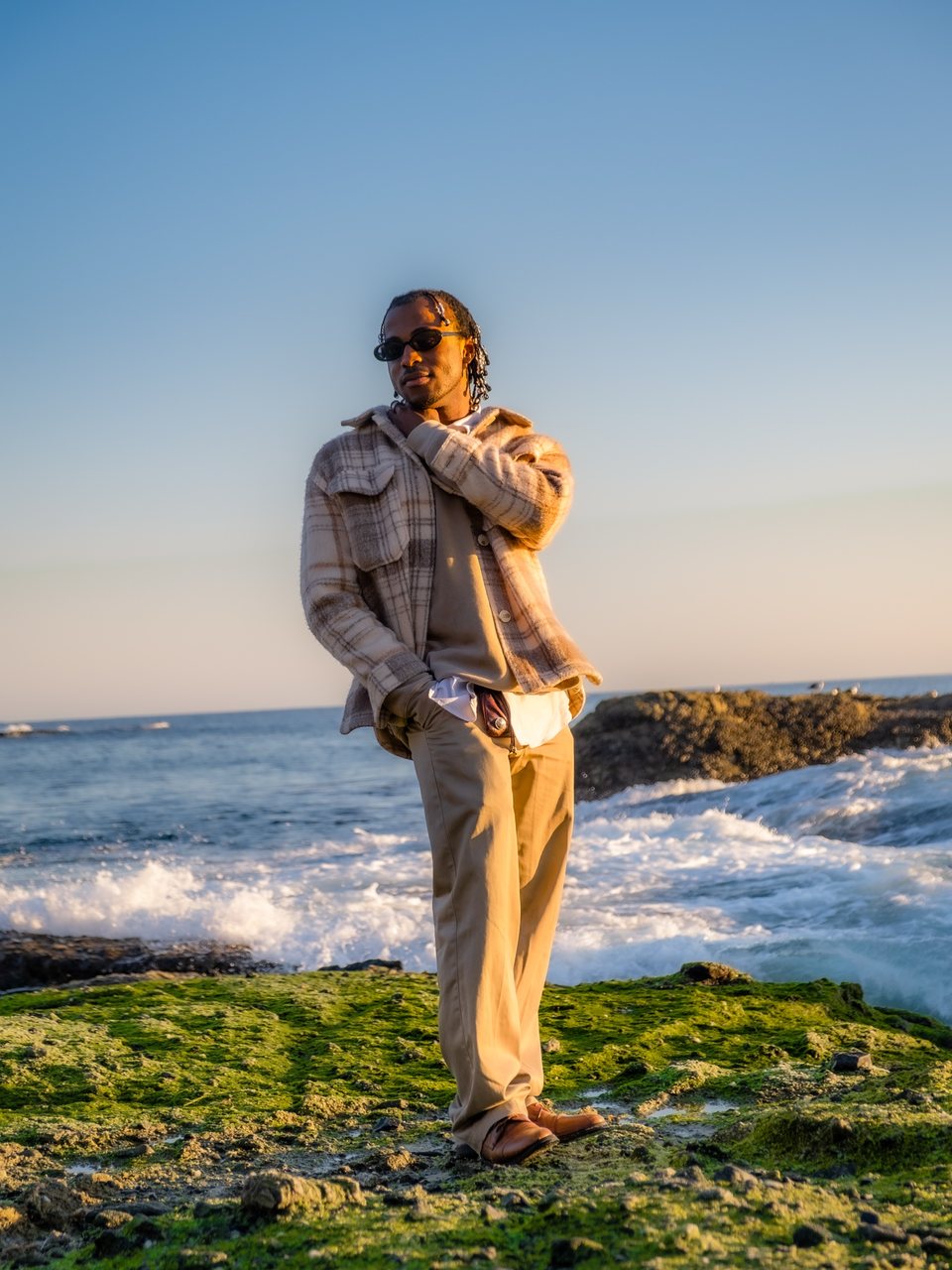 A man in a plaid shirt jacket and sunglasses standing on mossy rocks by the sea at golden hour