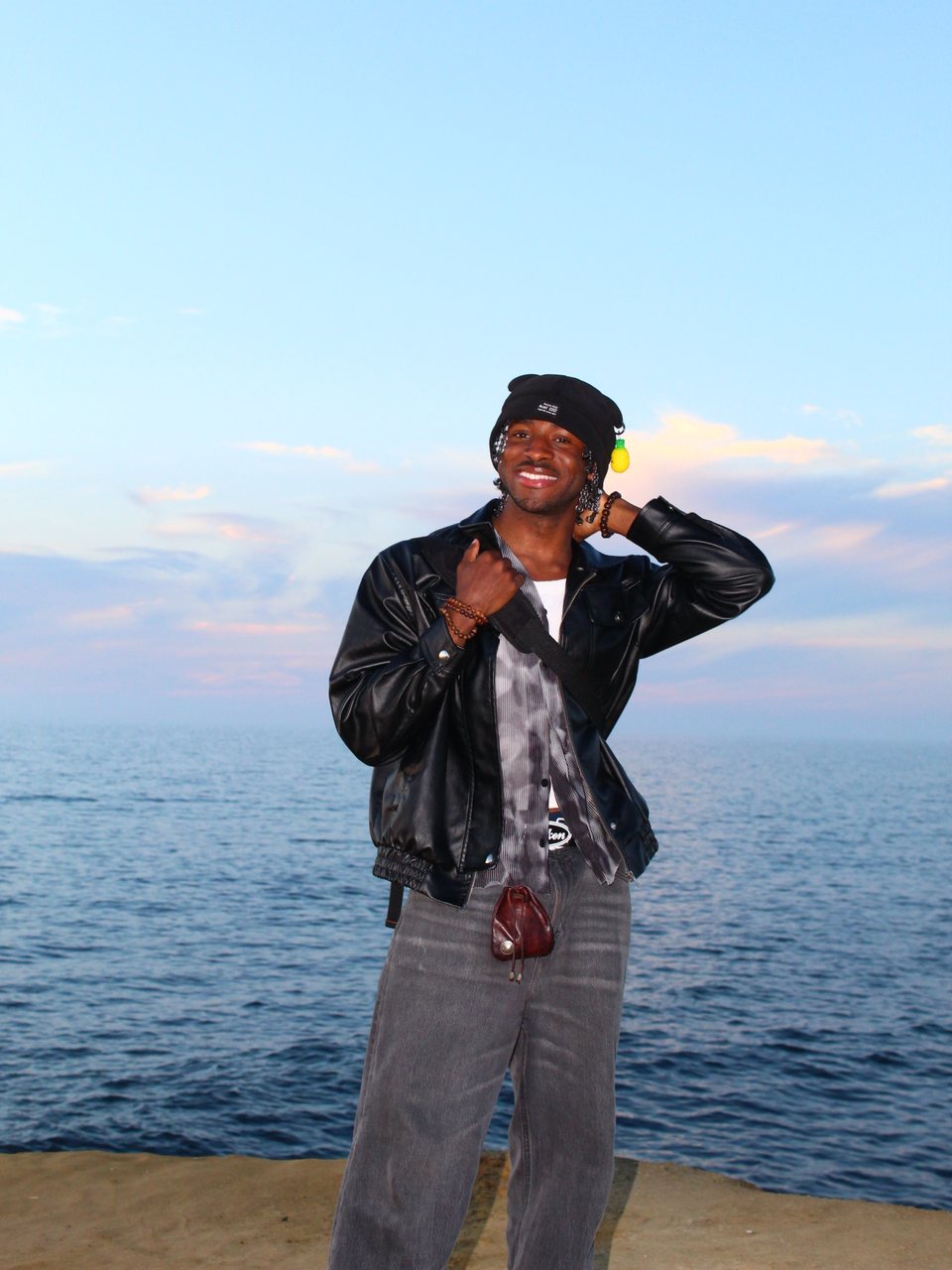 A smiling man in a leather jacket and beanie on the shore, the ocean behind him